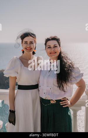 Femmes amis terrasse deux dames élégantes dans des tenues blanches vintage souriant sur un balcon côtier ensoleillé pendant l'heure dorée Banque D'Images