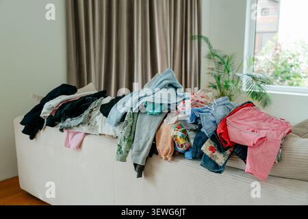 Une grande pile désordonnée de vêtements et de vêtements colorés pour bébé empilés sur un canapé lumineux dans un salon, attendant d'être pliés ou repassés. Photo de stock Banque D'Images