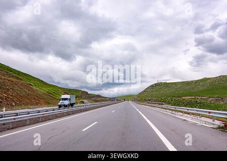 Large autoroute s'étend à travers des collines verdoyantes sous le ciel nuageux. Un seul camion blanc avance Banque D'Images