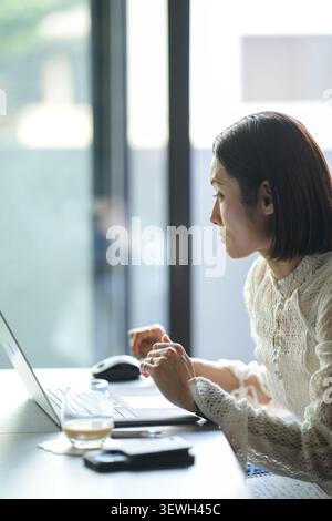 Jeune femme assise au bureau, concentrée sur l'écran d'ordinateur portable. La lumière douce filtre à travers la fenêtre derrière elle. Elle porte un pull délicat et texturé. Repos des lunettes b Banque D'Images