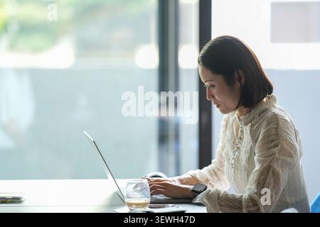 Une lumière douce brille à travers les grandes fenêtres. Femme s'assoit tranquillement, concentré sur l'écran de l'ordinateur portable. Ses doigts se déplacent doucement sur les touches du clavier. Le verre à café reste n Banque D'Images