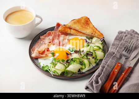 Petit déjeuner continental - œufs, bacon, micro-légumes verts et tranches de concombre sur une assiette sur la table. Petit déjeuner sain. Banque D'Images