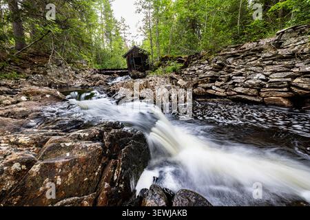 L'eau se précipite sur des rochers déchiquetés, créant des cascades blanches mousseuses. Les murs de pierre robustes encadrent l'énergie sauvage du ruisseau. Une forêt vert profond se dessine beh Banque D'Images
