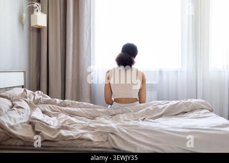 Jeune femme afro-américaine assise sur le bord du lit dans la chambre, le dos tourné, regardant par la fenêtre lumineuse. Scène de style de vie matinale calme à la maison Banque D'Images