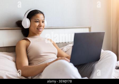 Femme noire heureuse assise dans le lit portant des écouteurs et utilisant un ordinateur portable Banque D'Images