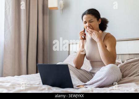 Femme noire assise sur le lit avec ordinateur portable, parlant sur smartphone et bâillant Banque D'Images