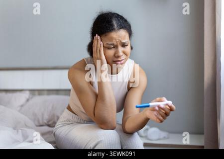 Femme noire assise sur le lit et regardant inquiet tout en tenant le test de grossesse Banque D'Images