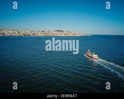 Un ferry orange et blanc navigue sur le Tage avec le paysage urbain de Lisbonne en arrière-plan. Cette image capture la vie quotidienne et le transit alon Banque D'Images