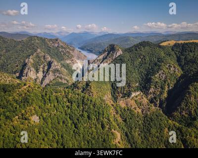 Une perspective aérienne révèle le barrage de Vidraru en béton retenant les eaux bleues du lac Vidraru, entouré de vastes montagnes boisées vertes et d Banque D'Images
