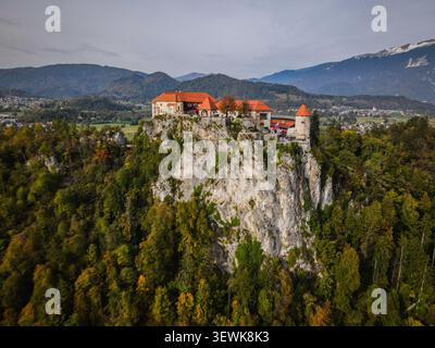 Bled Castle avec ses toits rouges se dresse au sommet d'une falaise rocheuse entourée d'arbres verts et oranges, avec des montagnes lointaines et une ville dans l'arrière-pays Banque D'Images