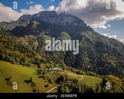 Une maison se trouve sur une colline verdoyante avec un chemin sinueux menant à elle, soutenu par une grande montagne couverte d'arbres changeant de couleur. Les Alpes juliennes sont un Banque D'Images