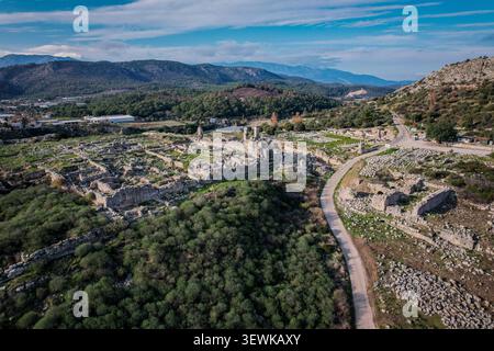 Une vue aérienne montre les vastes ruines de Patara et une route sinueuse à travers des collines verdoyantes et des montagnes sous un ciel bleu. Ce site historique est un signi Banque D'Images