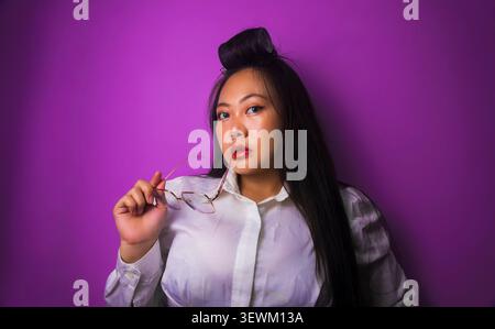 Jeune femme asiatique portant une blouse blanche tient des lunettes tout en posant sur un fond de studio violet avec espace de copie Banque D'Images