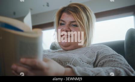 Une femme est confortablement assise à l'intérieur lisant un livre avec une expression concentrée sur son visage. Banque D'Images