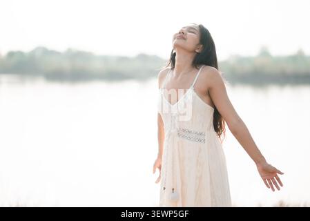 Femme asiatique en robe de dentelle blanche debout par un lac lumineux avec les yeux fermés et les bras ouverts, montrant la liberté, la paix, la respiration, la pleine conscience, la nature bien Banque D'Images