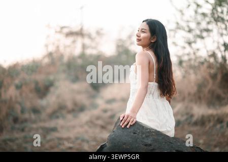 Vue de côté de femme asiatique en robe de dentelle blanche assise sur un rocher sombre à l'extérieur, souriant doucement dans la nature paisible, montrant la beauté, la solitude calme, relaxa Banque D'Images