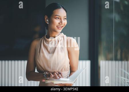 Femme d'affaires asiatique souriante à l'aide d'une calculatrice dans un cahier près de la fenêtre en verre, montrant la planification budgétaire, les notes comptables, la finance mobile, productivit Banque D'Images