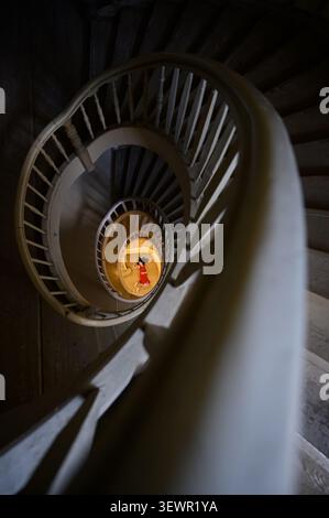 Vue en angle élevé d'un escalier en colimaçon vintage en bois avec une belle femme dans une robe rouge couchée au centre du plancher inférieur éclairé Banque D'Images