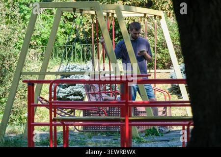 KIEV, UKRAINE - 28 JUIN 2026 : homme regardant son smartphone tout en se tenant debout près d'un enfant sur une aire de jeux dans le jardin botanique national M. M. Gryshko Banque D'Images
