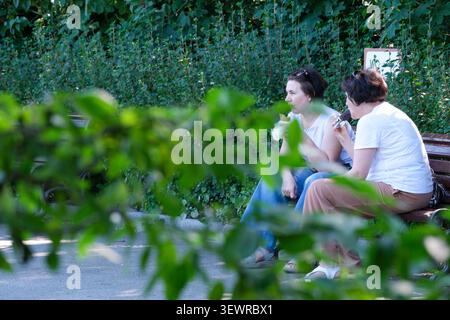 KIEV, UKRAINE - 28 JUIN 2026 : deux femmes dégustant de la crème glacée assis sur un banc de parc dans le jardin botanique national M. M. Gryshko par un été chaud Banque D'Images