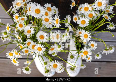 Bouquets de Marguerites blanches en bottes en caoutchouc blanc. Vue aérienne de marguerites blanches fraîches avec des centres dorés disposés dans des bottes en caoutchouc blanc sur bois gris Banque D'Images