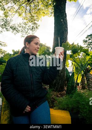 Femme tient une tasse de café. Elle porte une veste-cache noire et un Jean bleu. De grands arbres et des plantes vertes l'entourent. Elle a l'air détendue et satisfaite. IDE Banque D'Images