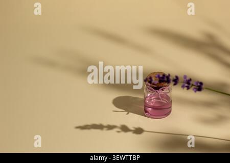 Composition de bien-être minimaliste avec des fleurs de lavande et une bouteille en verre rose avec des ombres naturelles douces sur un fond beige chaud, créant un CA Banque D'Images