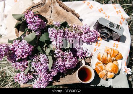 Une scène de pique-nique printanière confortable avec un grand bouquet de lilas violets, une tasse de thé, des croissants frais, et une caméra de film vintage sur une surface en bois, Banque D'Images