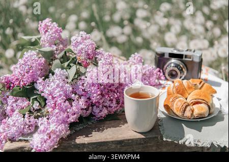 Une scène de pique-nique printanière confortable avec un grand bouquet de lilas violets, une tasse de thé, des croissants frais, et une caméra de film vintage sur une surface en bois, Banque D'Images