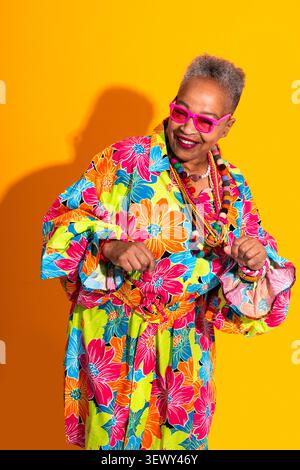 Femme senior noire portant un portrait de vêtements modernes sur fond - femme senior portant une robe florale colorée, des lunettes de soleil roses et un collier de perles, Banque D'Images