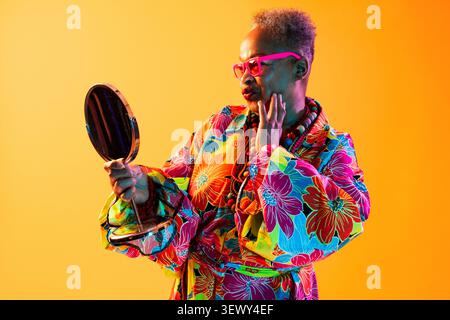 Femme senior noire portant un portrait de vêtements modernes sur fond - femme senior en robe florale colorée et lunettes roses regardant un petit miroir, t Banque D'Images