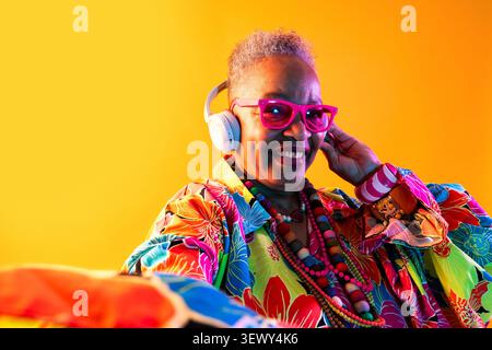Femme senior noire portant un portrait de vêtements modernes sur fond - femme senior souriant, portant une chemise florale vibrante, des lunettes roses et des écouteurs A. Banque D'Images