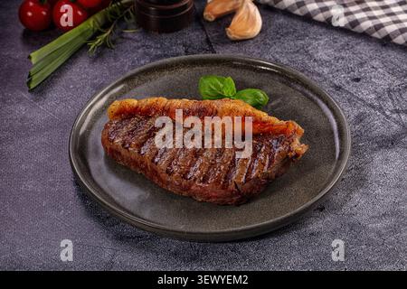 Bifteck picanha grillé, chapeau d'aloyau supérieur, servi sur une assiette rustique gris foncé. Garni de basilic frais. Le fond montre les tomates et l'ail pour saler, Banque D'Images