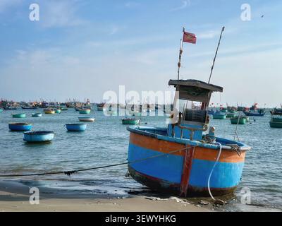 Bateau à panier rond coloré traditionnel sur une plage vietnamienne. Un bateau à panier rond traditionnel bleu et orange vif repose sur une rive sablonneuse. Banque D'Images