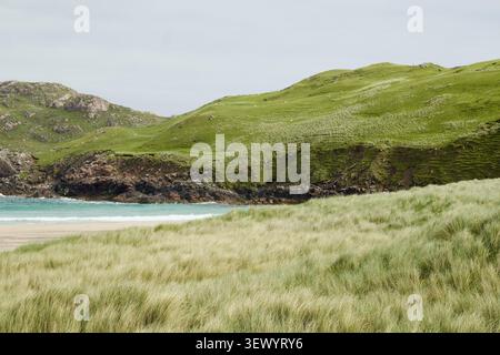 L'herbe des dunes balayée par le vent guide le regard vers une baie turquoise encadrée de collines vertes à Cliff Beach, île de Lewis, Hébrides extérieures, Écosse Banque D'Images