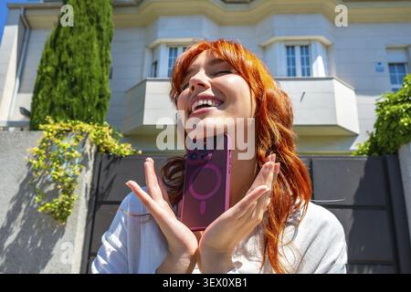 Femme rousse souriant les yeux fermés, tenant un smartphone dans un étui magenta doté d'un anneau magnétique, représentant le style de vie moderne et la technologie Banque D'Images