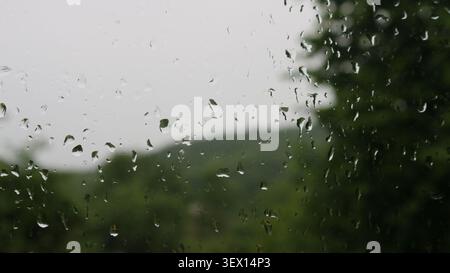 Grandes gouttes de pluie sur une vitre. À travers la surface humide, une silhouette très floue d'un paysage verdoyant avec des collines et des arbres sous un ciel couvert Banque D'Images