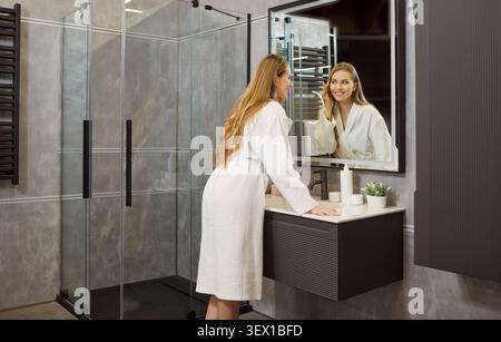 Femme dans la salle de bain moderne, debout regardant le miroir après la douche, le bain, faire des soins de beauté Banque D'Images