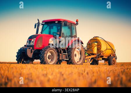 Tracteur agricole rouge avec remorque de réservoir jaune dans le champ de blé au coucher du soleil Banque D'Images