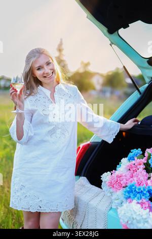 Femme blonde assise dans le coffre ouvert d'une voiture compacte avec des hortensias colorées et des verres à vin lors d'un pique-nique au coucher du soleil Banque D'Images