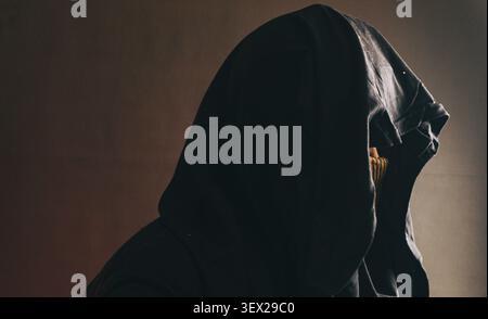 Un personnage masculin mystérieux se tient de profil portant une cagoule lourde sombre et un masque jaunâtre texturé, créant une atmosphère profondément secrète. Drame doux Banque D'Images