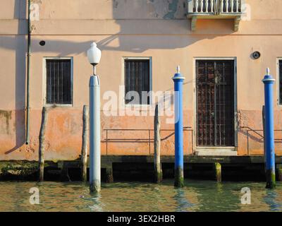 Façade de bâtiment altérée le long du canal de Venise avec des poteaux d'amarrage colorés et des fenêtres barrées Banque D'Images