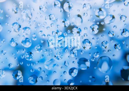 Des gouttelettes d'eau bleues s'accrochent à une surface transparente avec des fleurs. Chaque goutte réfracte la lumière, créant des reflets vibrants. L'arrière-plan s'illumine avec du Sof Banque D'Images
