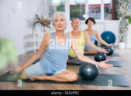 Vieille femme pratiquant la pose demi-lotus avec softball sous le bras dans la salle de fitness légère Banque D'Images