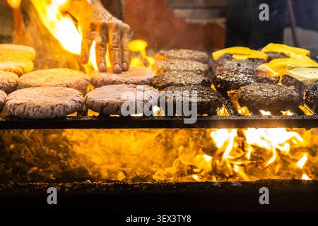 Gros plan sur les mains d'un chef disposant des galettes de viande crues et cuites sur une grille chaude avec des flammes rugissantes en dessous, préparant un hamburg artisanal traditionnel Banque D'Images
