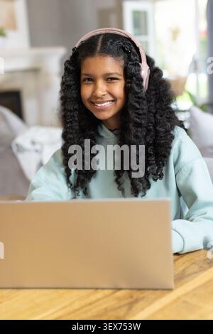Jeune fille afro-américaine étudiant sur ordinateur portable à la maison portant un sweat à capuche à la menthe, écouteurs roses Banque D'Images