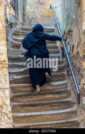 Femme en abaya noire gravissant des marches en pierre usées dans une ruelle du Caire, Égypte Banque D'Images