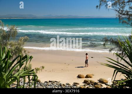 Australie, Nouvelle-Galles du Sud, Byron Bay, Wategos Beach, plage de sable bordée par l'océan Pacifique, avec un marcheur sur le rivage, vagues, rochers, végétation côtière et collines de l'arrière-pays en arrière-plan Banque D'Images