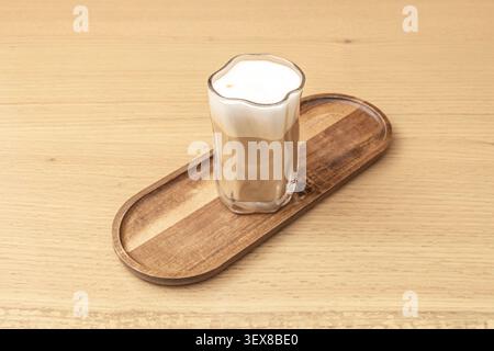 Latte en couches avec une épaisse mousse de lait blanche servi dans un gobelet en verre ondulé unique reposant sur un plateau en bois étroit au sommet d'une table lumineuse Banque D'Images
