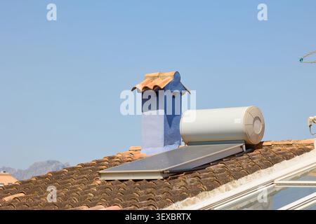 Vue rapprochée d'un système de chauffe-eau solaire monté sur un toit traditionnel en tuiles d'argile à côté d'une cheminée bleue sous un ciel bleu clair à Denia, en Espagne Banque D'Images